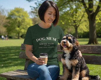 May include: A dark green short-sleeved t-shirt featuring the white text DOG MUM SOCIAL CLUB EST. 2024 WALKS COFFEE MUDDY PAWS. The shirt is paired with blue denim jeans, and a black, tan, and white tri-color dog sits beside the wearer.