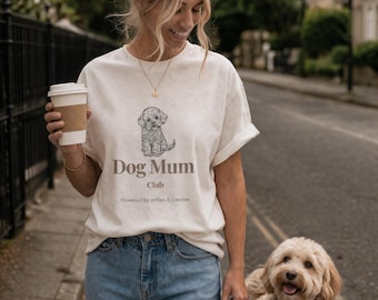 May include: An off-white short-sleeve t-shirt featuring a line-art illustration of a small dog above the text "Dog Mum Club" and "Powered by coffee & cuddles" in a brown sans-serif font, paired with light blue denim jeans.