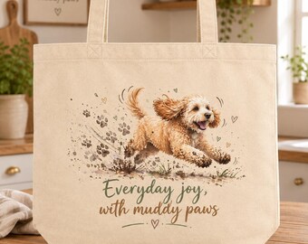 May include: A beige canvas tote bag featuring a watercolor illustration of a golden-colored dog running through mud with paw prints. The text "Everyday joy, with muddy paws" is printed below the dog in dark green and brown script lettering.