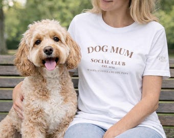 May include: A white short-sleeved t-shirt featuring brown text that reads DOG MUM SOCIAL CLUB EST. 2024 WALKS COFFEE MUDDY PAWS on the chest and a small script logo on the sleeve, paired with light blue denim jeans and a fluffy tan dog.