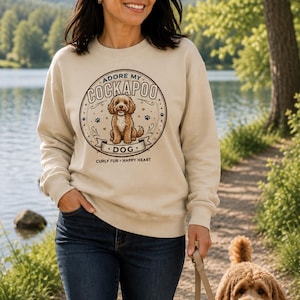May include: A beige crewneck sweatshirt featuring a circular graphic with a brown Cockapoo dog illustration. The text reads ADORE MY COCKAPOO DOG and CURLY FUR HAPPY HEART. The design includes small paw prints and decorative flourishes in a dark brown ink.