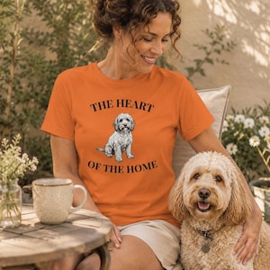 May include: An orange short-sleeved t-shirt featuring a graphic of a small grey dog sitting in the centre. The text "THE HEART OF THE HOME" is printed in black capital letters above and below the dog illustration on the front of the shirt.