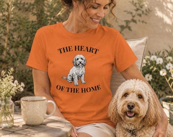 May include: An orange short-sleeved t-shirt featuring a graphic of a small gray dog sitting in the center. The text "THE HEART OF THE HOME" is printed in black capital letters above and below the dog illustration on the front of the shirt.