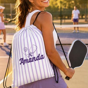 May include: A white drawstring backpack featuring vertical light purple stripes, a graphic illustration of two pickleball paddles, and the name Amanda printed in a bold purple script font across the center of the bag.