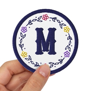 May include: A circular embroidered patch featuring a dark blue border and a white centre. The letter M is stitched in dark blue in the middle, surrounded by a decorative wreath of small red, yellow, and purple flowers with green leaves.