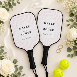 Könnte beinhalten: Zwei Pickleball-Schläger mit schwarzen Griffen und weißen Schlagflächen mit einem rosa geometrischen Linienmuster. Der Text lautet GAYLE and ROGER May 20, 2027. Die Schläger sind umgeben von weißen Rosen, Eukalyptusblättern, zwei Goldringen und zwei gelben Bällen.