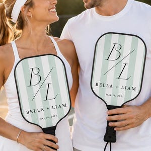 Könnte beinhalten: Zwei personalisierte Pickleball-Schläger mit einem hellgrün-weißen vertikalen Streifenmuster. Jeder Schläger zeigt das Monogramm B/L, die Namen BELLA + LIAM und das Datum May 20, 2027 in schwarzer Schrift mit schwarzem Griff und Handschlaufe.