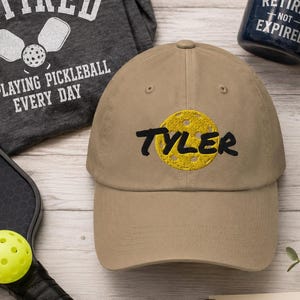 以下が含まれることがあります： 黄色のピックルボールのグラフィックと黒い文字でTylerと書かれたベージュの野球帽。セットには、ピックルボールのテキストが入ったダークグレーのTシャツ、Retired Not Expiredと書かれた紺色のマグカップ、金属製のボトル、ピックルボールのラケット、ボールが含まれています。