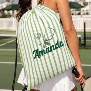 May include: A drawstring backpack featuring a white and light green vertical striped pattern. The bag displays a dark green graphic of a pickleball paddle and ball, with the name Amanda printed in a dark green cursive font across the center.