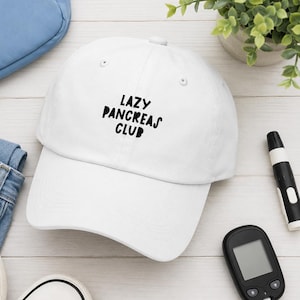May include: A white baseball cap with the black text "LAZY PANCREAS CLUB" printed on the front. The cap features a structured crown, a curved brim, and a button on top. The design is centered on the front panel in a bold, sans-serif font.