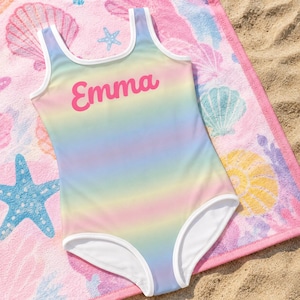 Può includere: Un costume da bagno intero con una sfumatura arcobaleno pastello e bordi bianchi lungo lo scollo e le aperture delle gambe. Il nome Emma è stampato in un carattere corsivo rosa audace al centro della zona del petto del capo.