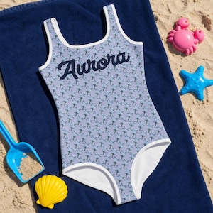 Può includere: Un costume da bagno intero con base color viola chiaro caratterizzato da un motivo ripetuto di piccoli fiori blu e steli verdi. La parola Aurora è stampata in corsivo blu scuro sul petto. Il costume ha bordi bianchi lungo lo scollo e le gambe.
