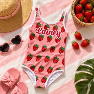 Può includere: Un costume da bagno intero rosa con un motivo a fragole ripetuto e il nome "Lainey" stampato in corsivo bianco sul petto. Il costume presenta un bordo bianco lungo lo scollo e le aperture per le braccia, con un design classico a girocollo.