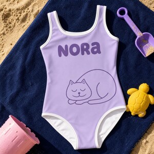 Può includere: Un costume da bagno intero color viola chiaro con bordini bianchi, caratterizzato dal nome NORA stampato in lettere viola scuro sopra un semplice disegno al tratto di un gatto che dorme, appoggiato su un asciugamano blu scuro.