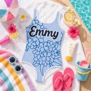 Può includere: Un costume da bagno intero azzurro con un motivo floreale blu scuro e il nome Emmy stampato in un audace carattere corsivo blu scuro sul petto. Il costume presenta uno scollo rotondo e bordi bianchi lungo le aperture delle braccia e il collo.