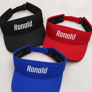May include: Three athletic visors in black, red, and blue, each featuring the name Ronald embroidered in white thread on the front panel. The visors include an adjustable strap with a black buckle and an interior band labelled with the text 110 FLEXFIT.