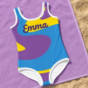 Può includere: Un costume da bagno intero con un vivace motivo astratto di forme organiche blu, viola e gialle. Il capo presenta un bordo bianco lungo lo scollo e le aperture delle gambe, con il nome Emma stampato in corsivo viola scuro sul petto.