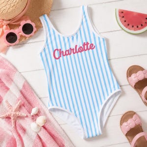 Può includere: Un costume da bagno intero a righe verticali azzurre e bianche con il nome Charlotte stampato in corsivo rosa sul petto. Il costume presenta uno scollo rotondo e bordi bianchi lungo le aperture delle braccia e il girocollo.