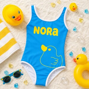Può includere: Un costume da bagno intero blu brillante con bordi bianchi, il nome NORA stampato in giallo e il disegno stilizzato di una papera di gomma gialla. Il costume è circondato da papere di gomma gialle, occhiali da sole blu e gemme colorate.