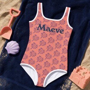 Può includere: Un costume da bagno intero color corallo aranciato con un motivo floreale ripetuto a linee blu scuro. Il capo presenta un bordo bianco lungo lo scollo e le aperture delle gambe, con il nome Maeve stampato in blu scuro sul petto.