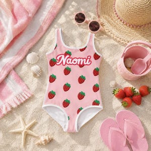 Può includere: Un costume da bagno intero rosa chiaro con un motivo ripetuto di fragole rosse e il nome Naomi stampato in corsivo bianco sul petto. Il costume presenta un bordo bianco lungo lo scollo e le aperture per le braccia.