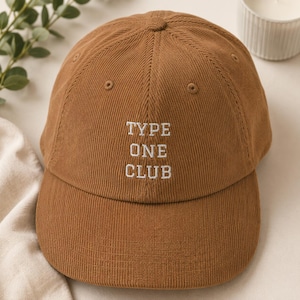 May include: A brown corduroy baseball cap featuring the embroidered white text "TYPE ONE CLUB" centered on the front panel. The hat has a structured crown, a curved brim, and stitched eyelets for ventilation, showcasing a classic six-panel design.