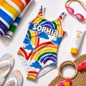 Può includere: Un costume da bagno intero con motivo arcobaleno, nuvole blu e il nome SOPHIA stampato in bianco. Il set include un asciugamano a righe arcobaleno, occhialini da nuoto rosa, un tubetto di crema solare SPF 50 e un paio di infradito.