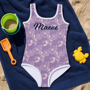 Può includere: Un costume da bagno intero color viola chiaro con un motivo ripetuto di contorni floreali rosa pallido e bordini bianchi. Il nome Maeve è stampato in corsivo blu scuro sul petto del capo, caratterizzato da uno scollo rotondo.