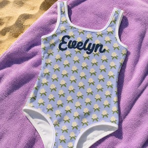 Può includere: Un costume da bagno intero color azzurro con un motivo ripetuto di piccoli fiori bianchi e foglie verdi. Il nome Evelyn è stampato in corsivo blu scuro sul petto. Il capo presenta bordi bianchi lungo lo scollo e le aperture per le gambe.