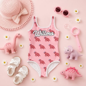 Può includere: Un costume da bagno intero rosa con un motivo ripetuto di dinosauri triceratopo rossi e il nome Willow in corsivo bianco. Gli accessori includono un cappello di paglia rosa, occhiali da sole rosa, sandali bianchi e tre piccole statuette di dinosauri.