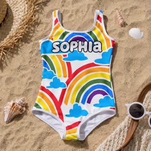 Può includere: Un costume da bagno intero con sfondo bianco e un motivo colorato di arcobaleni e nuvole blu. Il nome SOPHIA è stampato in grandi lettere bianche con contorno nero sulla parte del petto del capo.
