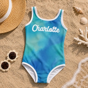 Può includere: Un costume da bagno intero con motivo tie-dye blu e verde acqua, caratterizzato da bordini bianchi lungo lo scollo e gli scalfi. Il nome Charlotte è stampato in corsivo bianco al centro del petto sul davanti del capo.