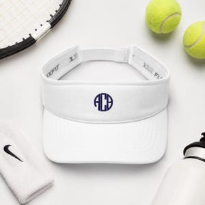 May include: A white visor featuring a dark blue circular monogram with the letters ACB embroidered on the front center. The visor has an adjustable strap at the back with the text FLEXFIT 110 visible on the inner band of the headwear.