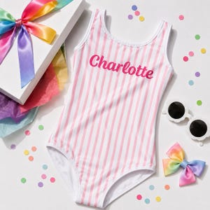 Può includere: Un costume da bagno intero senza maniche con righe verticali rosa chiaro e bianche. La parte anteriore del capo presenta il nome Charlotte stampato in un audace carattere corsivo rosa scuro. Il costume è presentato accanto a un nastro arcobaleno.