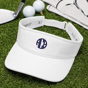 May include: A white golf visor featuring a navy blue circular monogram with the letters ACB embroidered on the front panel. The visor has an adjustable strap at the back with the text Flexfit 110 visible on the interior band of the headwear.