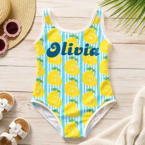 Può includere: Un costume da bagno intero senza maniche con un motivo di limoni gialli e foglie verdi su righe verticali azzurre e bianche. Il nome Olivia è stampato al centro in un carattere corsivo retro di colore blu scuro.