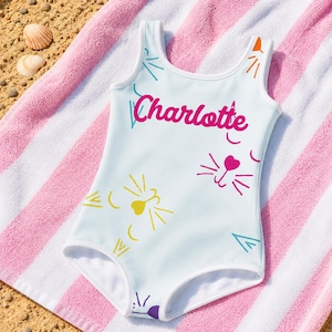 Può includere: Un costume da bagno intero bianco con il nome Charlotte scritto in corsivo rosa brillante. Il design presenta grafiche colorate di musi di gatto in giallo, rosa e viola, insieme a piccole forme astratte color ottanio e arancione sul davanti.