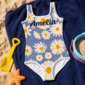 Può includere: Un costume da bagno intero con sfondo blu, motivo floreale a margherite bianche e il nome Amelia stampato in corsivo blu scuro sul petto. Il capo presenta uno scollo rotondo e bordi bianchi lungo gli scalfi e lo scollo.