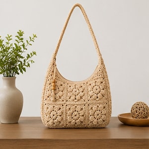 Peut inclure: Un sac à bandoulière beige en crochet avec un motif floral. Le sac a une forme incurvée, une longue bandoulière et un pompon avec des perles en bois. Le sac est posé sur une surface en bois.