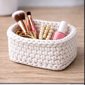 Könnte beinhalten: Ein kleiner, cremefarbener, gehäkelter Korb mit Beauty-Accessoires. Er enthält Make-up-Pinsel, goldfarbene Büroklammern, eine Kette aus Kunstperlen und eine Flasche rosa Nagellack. Der Korb steht auf einer hellen Holzoberfläche.