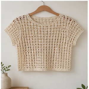 May include: A cream-colored, short-sleeved crochet top. The top is made with a loose, open weave, and hangs on a wooden hanger. The neckline and sleeves have a more tightly woven edge. The top is a neutral color and appears to be handmade.