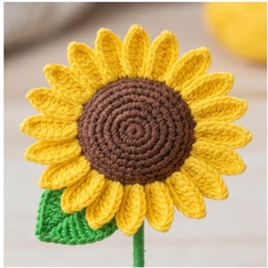 May include: A handmade crochet sunflower with yellow petals, a brown center, and a green leaf. The sunflower is on a green stem, creating a cheerful, decorative item. The craftwork showcases a detailed, textured design.