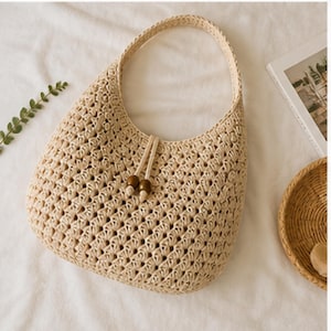 May include: A beige, open-weave crochet handbag with a rounded shape and a single handle. The bag features a drawstring closure with two wooden beads. The bag is made of a natural fiber material.