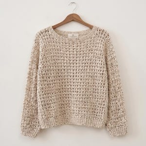 Puede incluir: Un suéter beige de manga larga con un patrón de rejilla de punto abierto y holgado y un cuello redondo. La prenda presenta una textura moteada en todo el tejido e incluye una pequeña etiqueta rectangular en el cuello interior que dice HEARTHEIDE.