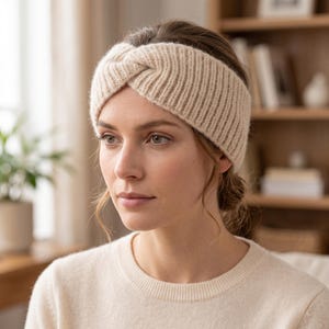 Può includere: Una fascia in maglia beige caratterizzata da una trama a coste e un dettaglio a nodo intrecciato centrale. La fascia è realizzata in un materiale di filato spesso e morbido che copre le orecchie e la fronte, offrendo una vestibilità aderente.