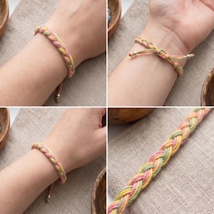 May include: A braided bracelet featuring interwoven strands of soft pink, pale yellow, and light green cord. The bracelet includes an adjustable sliding knot closure with small metallic beads at the ends of the loose cords for a secure fit.