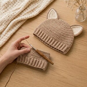 May include: A light brown crocheted hat with two pointed ears featuring cream-coloured inner details. Beside the hat is a partially completed rectangular crocheted piece in the same light brown yarn, being worked with a wooden crochet hook.