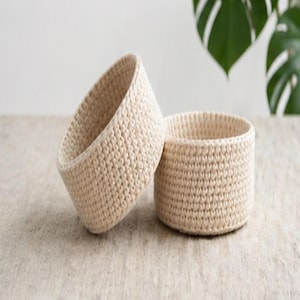 May include: Two handmade, off-white crochet baskets. One basket is upright, and the other is tilted, revealing the interior. The baskets are made of a thick, textured yarn and sit on a neutral surface. The baskets are round and have a simple, minimalist design.
