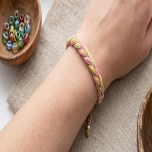 Könnte beinhalten: Geflochtenes Armband in Pastellfarben aus Rosa, Gelb und Grün. Das Armband besteht aus gewebten Fäden und hat einen verstellbaren Verschluss. Das Armband wird am Handgelenk getragen.