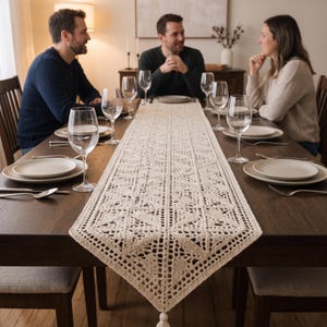 Puede incluir: Un camino de mesa rectangular de ganchillo en color crema con un intrincado patrón geométrico, detalles calados y un extremo puntiagudo terminado con una borla, centrado sobre una mesa de comedor de madera oscura con platos y copas.
